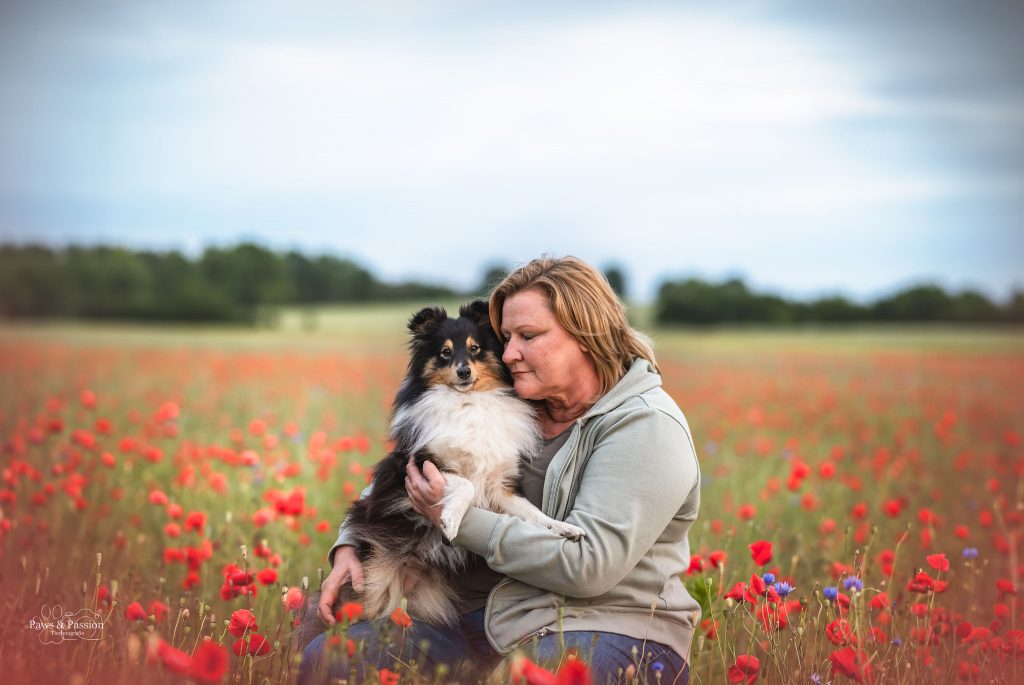 Das Bild zeigt eine Frau, die im Mohnfeld sitzt und ihre Hündin auf dem Arm hat und sich an sie schmiegt.
